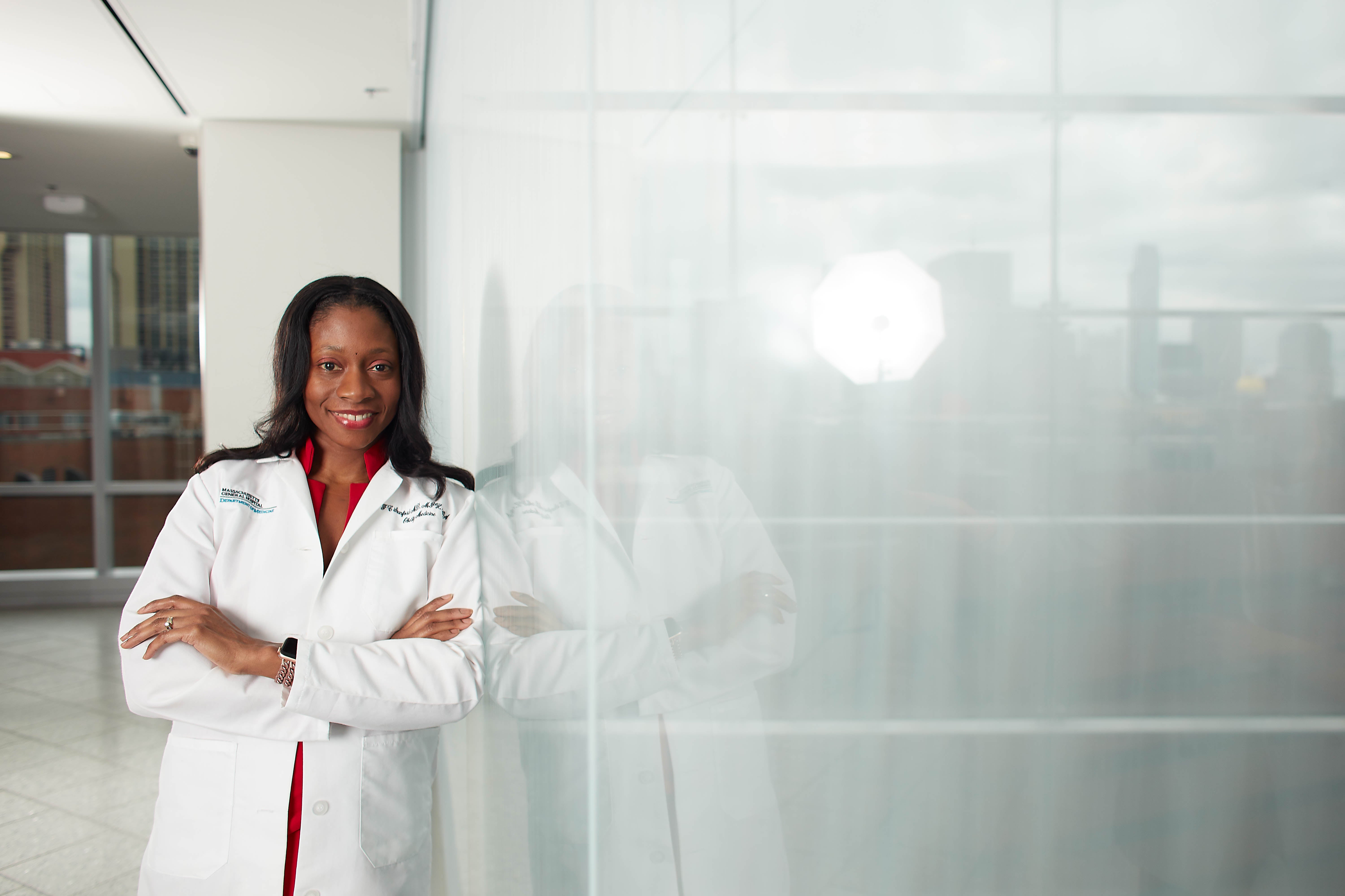 Fatima Cody Stanford in lab coat in white office hallway standing with arms crossed and leaning on reflective glass wall.