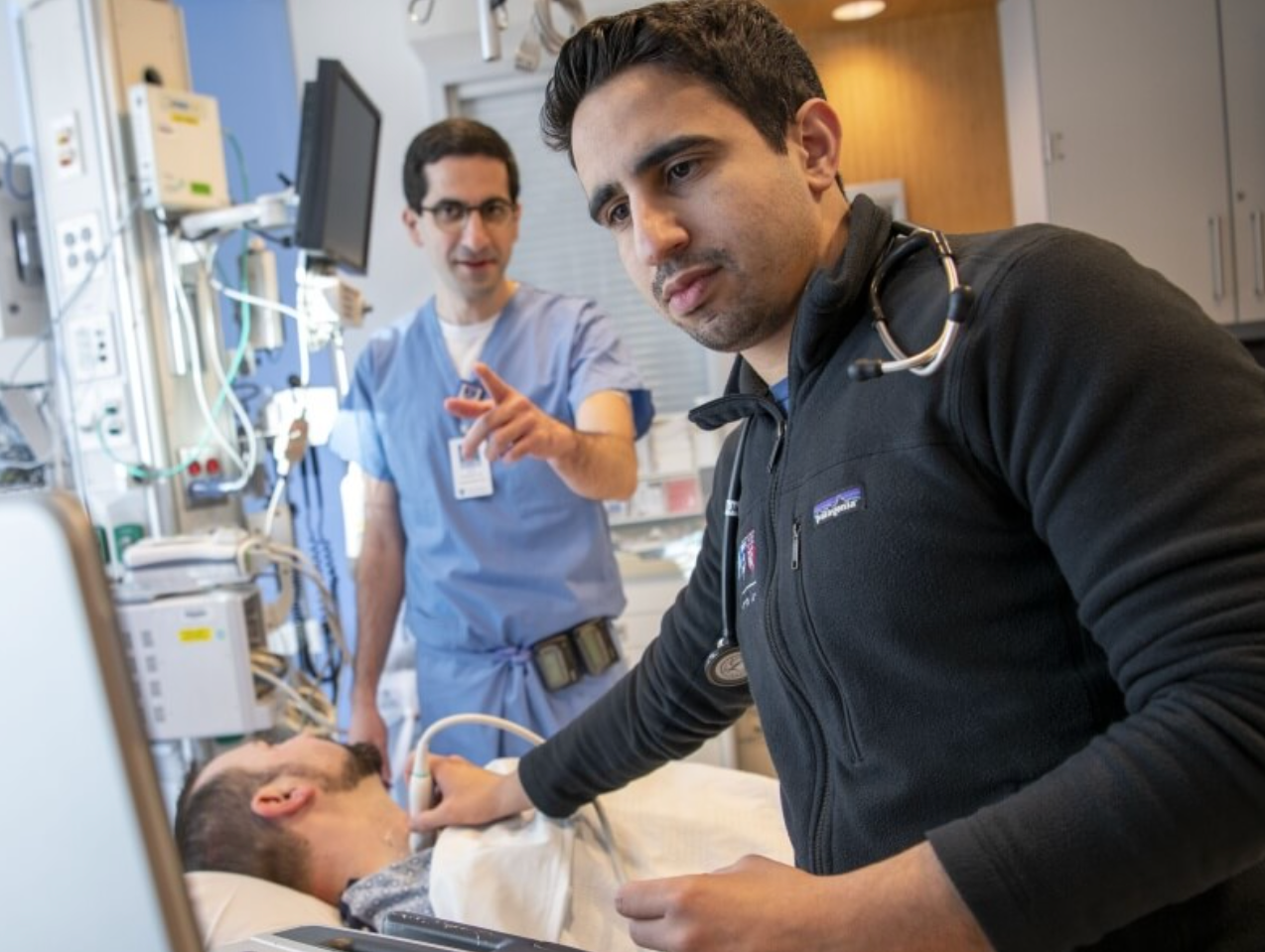 Haitham Alabsi looks at monitor while checking patients vitals in hospital room while another medical staff member in scrubs points at the monitor.