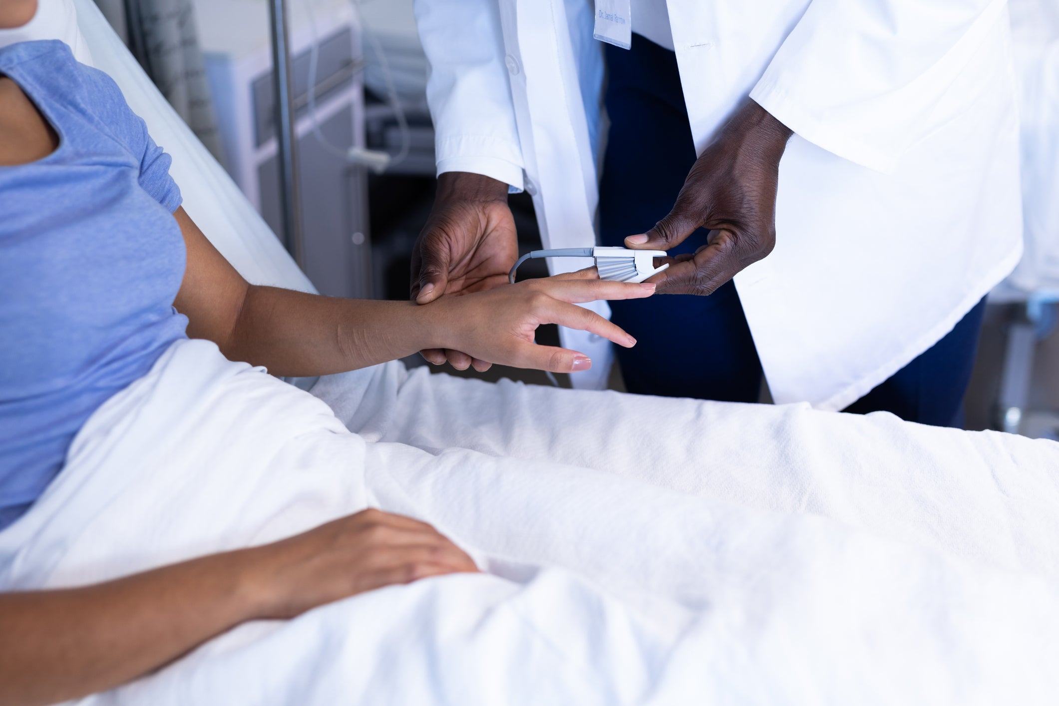 Midsection of African American male doctor putting oximeter on finger of woman in hospital bed.