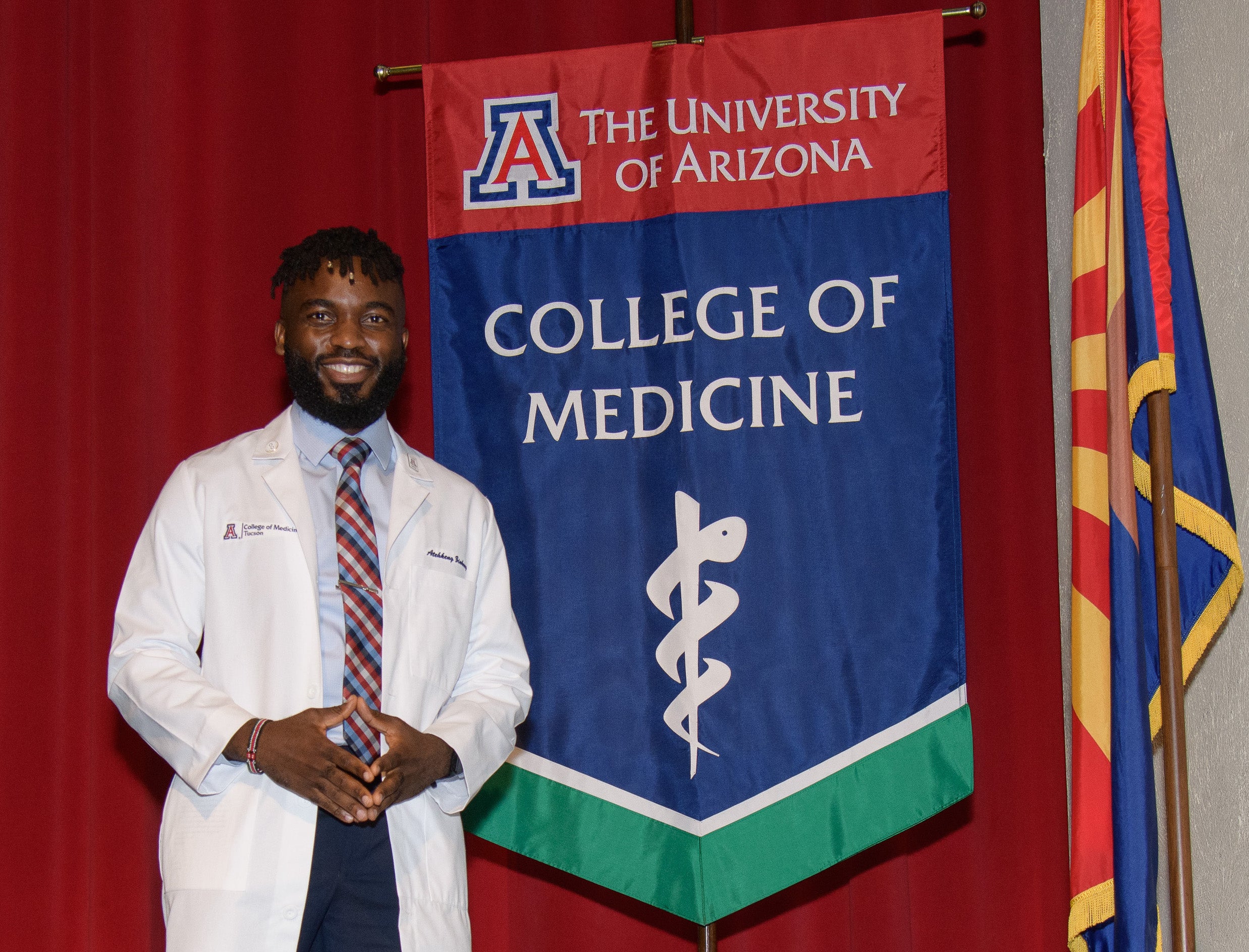 Ateh Zinkeng in his medical white coat standing next to The University of Arizona College of Medicine flag.